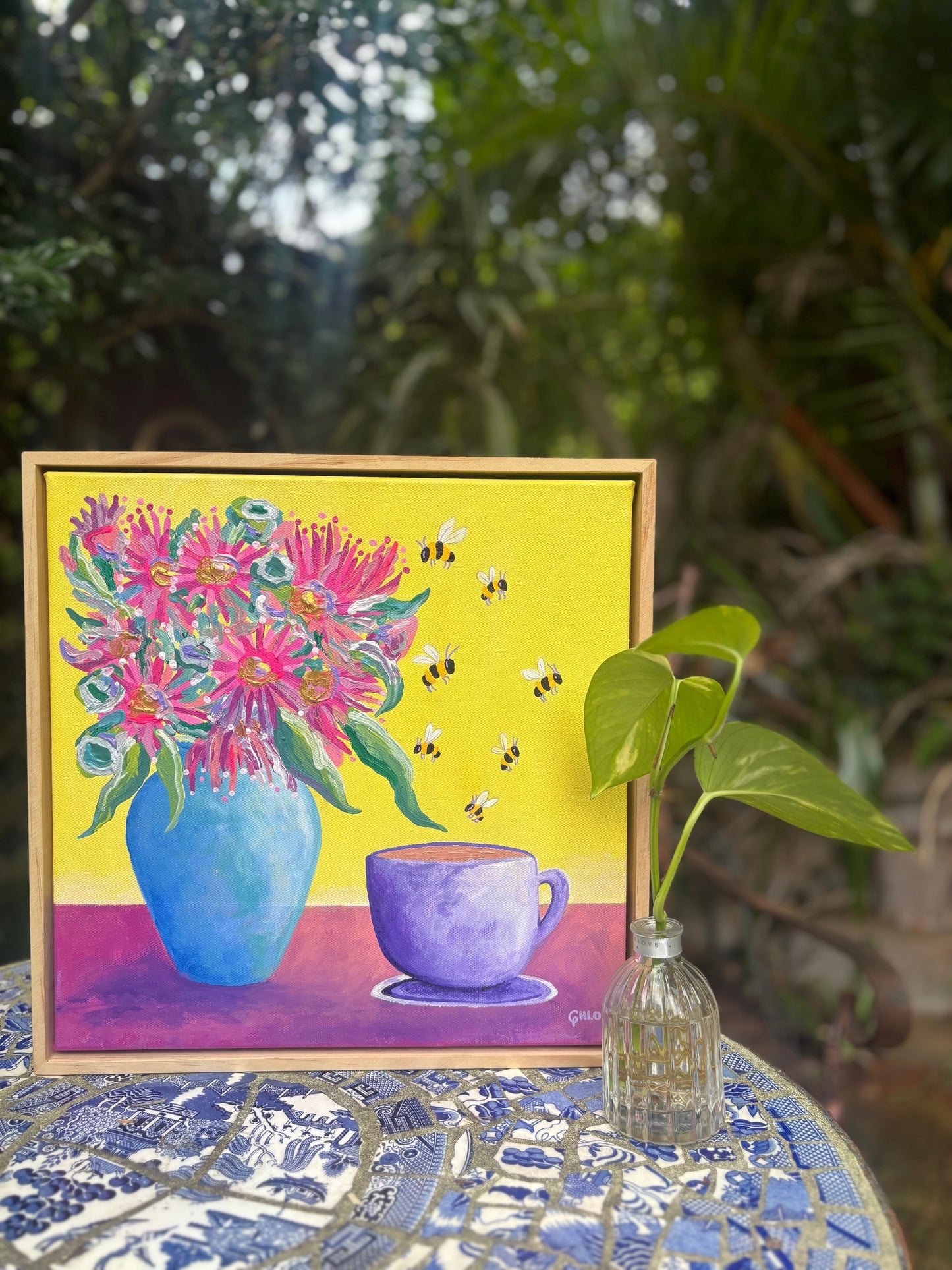 Colorful painting of flowers and a cup on a yellow background, with a plant in the foreground.