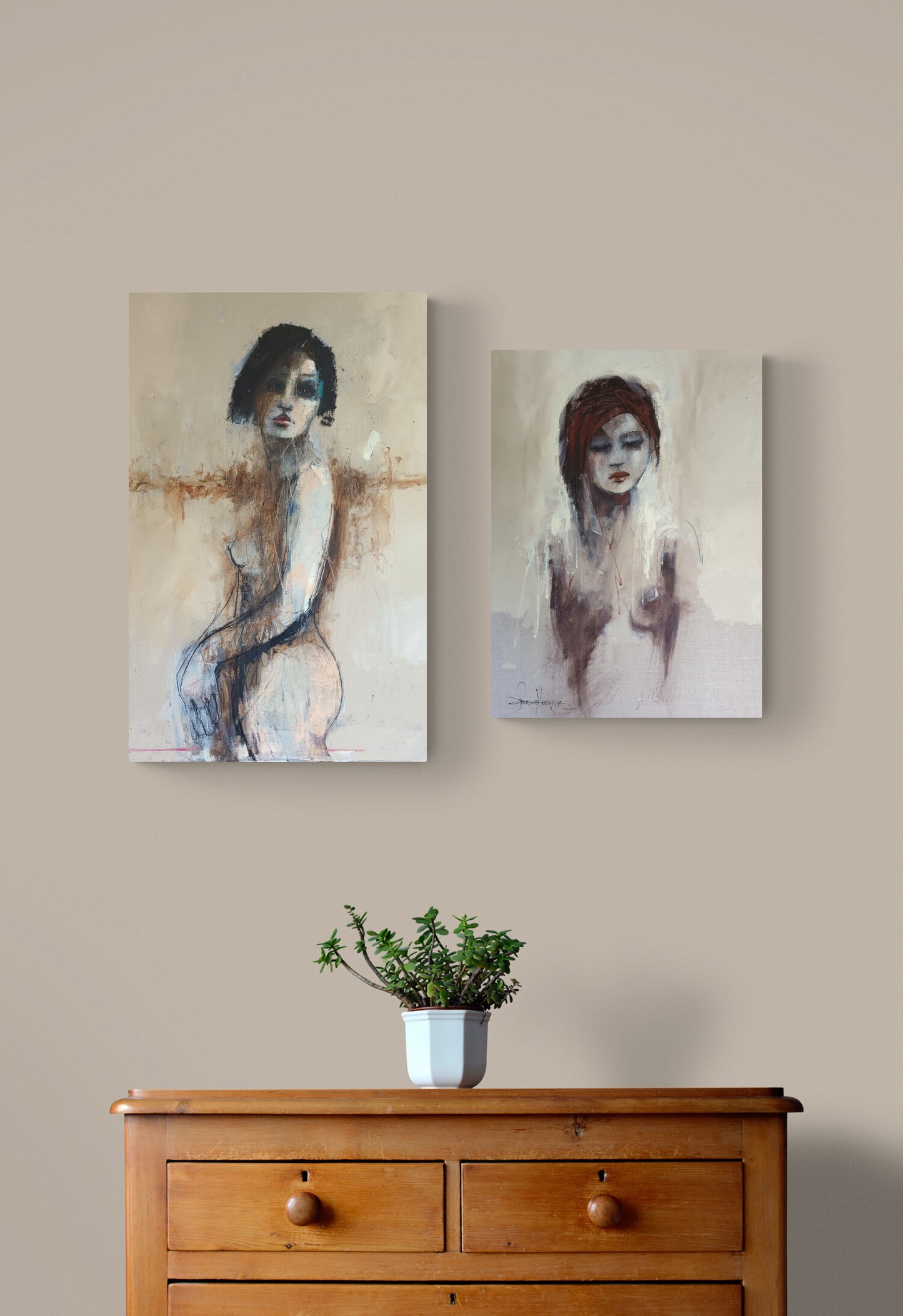 Two vertical canvas art prints depicting abstract female figures, displayed side-by-side on a beige wall above a wooden chest of drawers with a small potted plant.