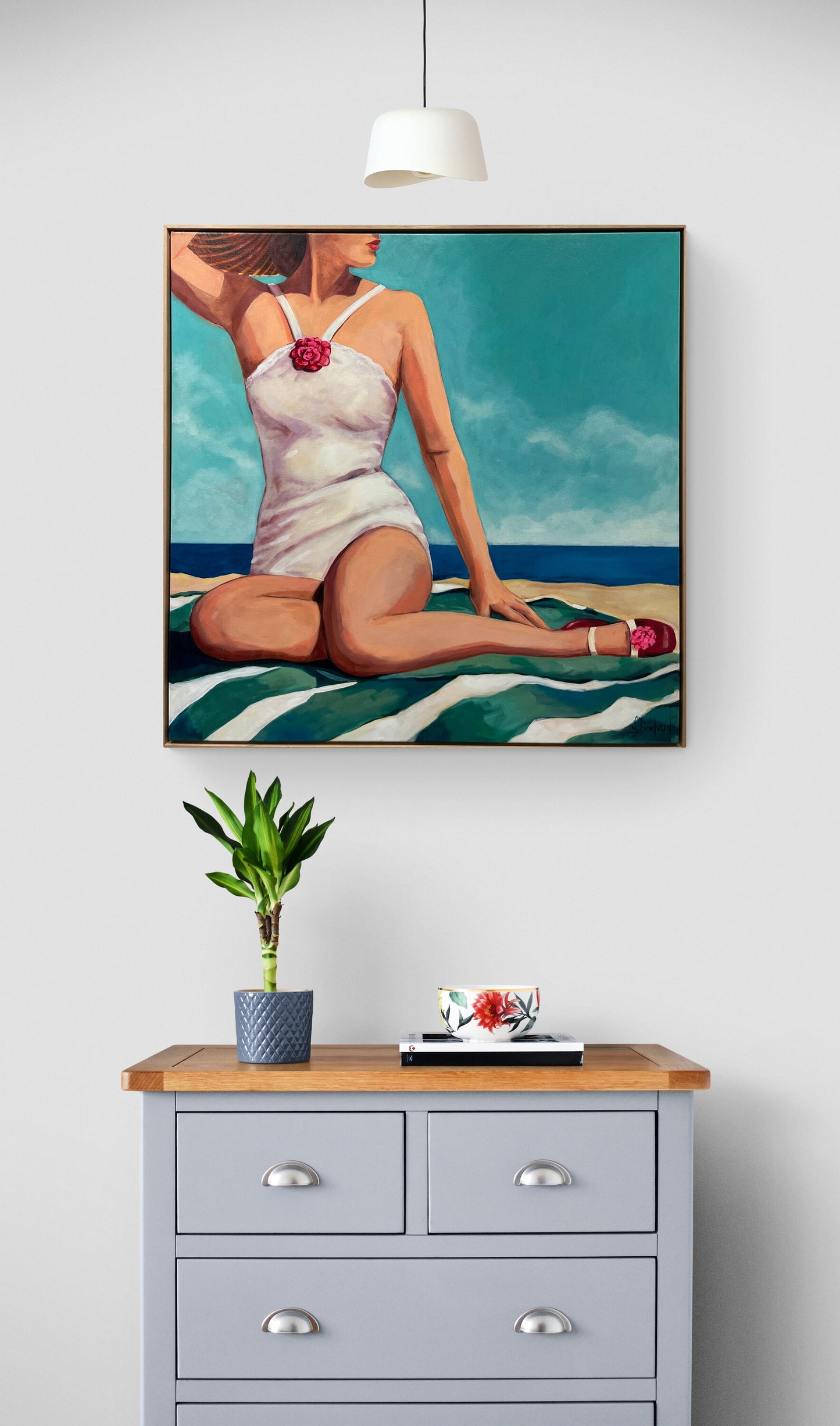 A framed art print depicting a retro-style woman in a white swimsuit on a striped beach towel, displayed on a light gray wall above a wooden dresser with a potted plant and a small book.