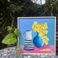 Framed painting of a blue vase with yellow flowers and a striped pitcher on a table outdoors.