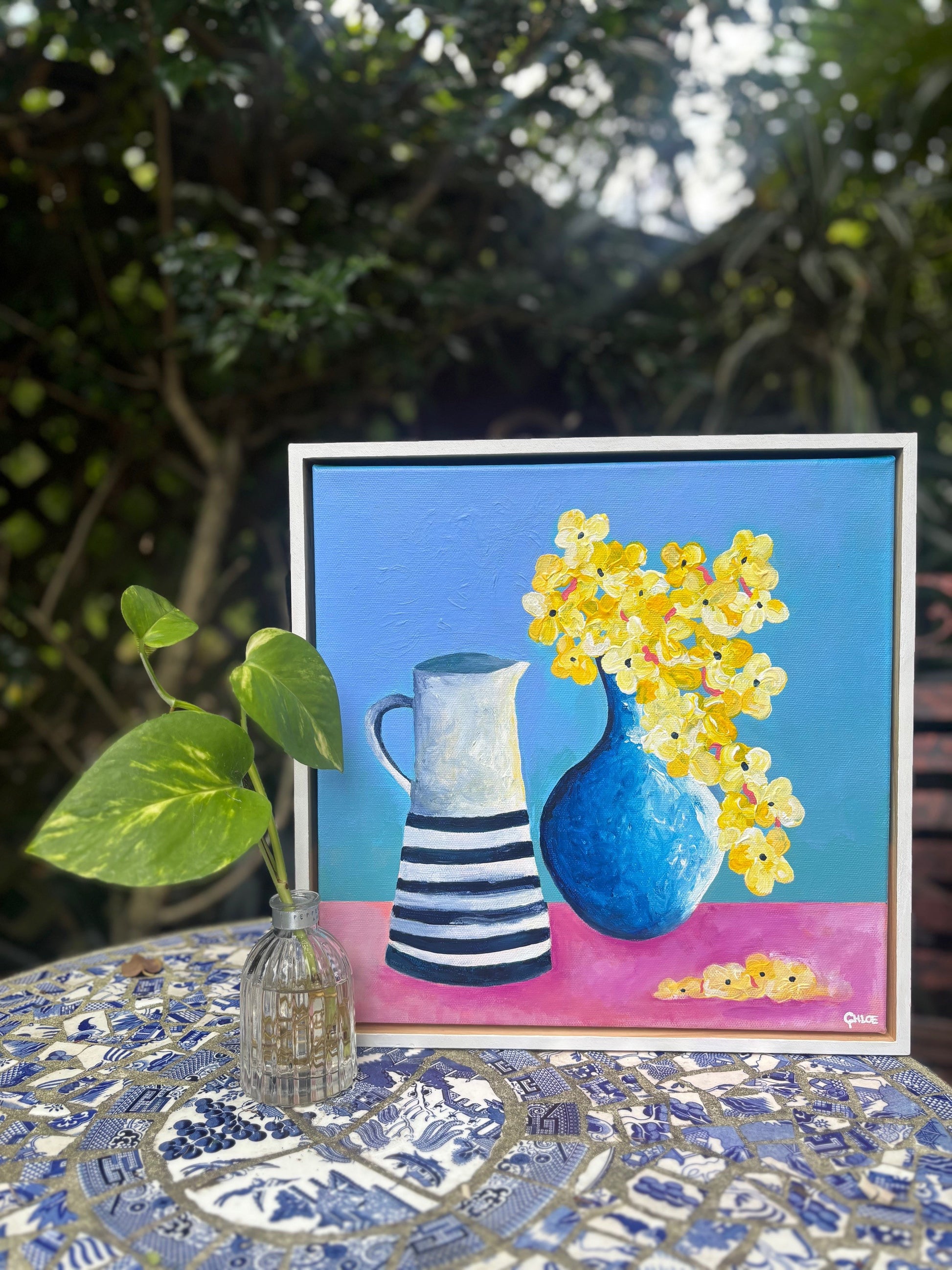Framed painting of a blue vase with yellow flowers and a striped pitcher on a table outdoors.