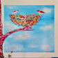 A colorful painting of a bird's nest on a tree branch against a blue sky with clouds, featuring two small birds, shown mounted on a beige wall.