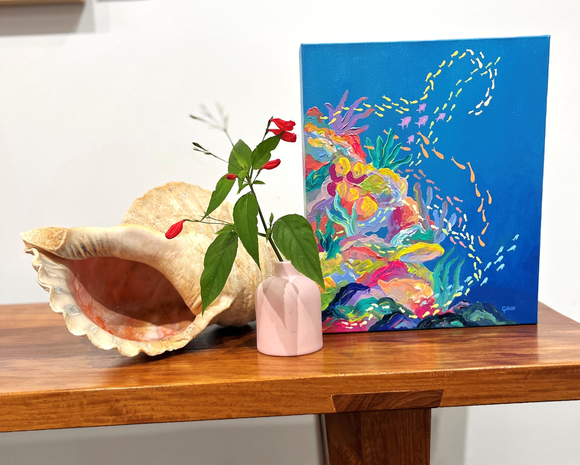 Colorful coral reef painting on a blue background with a large shell and pink vase on a wooden surface.