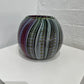 A hand-blown glass vase with a striped pattern of purple, green, gray, and gold, displayed on a white geometric platform against a white brick wall.