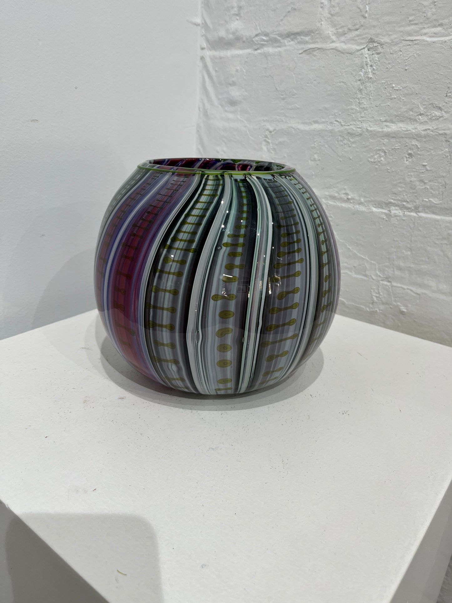 A hand-blown glass vase with a striped pattern of purple, green, gray, and gold, displayed on a white geometric platform against a white brick wall.