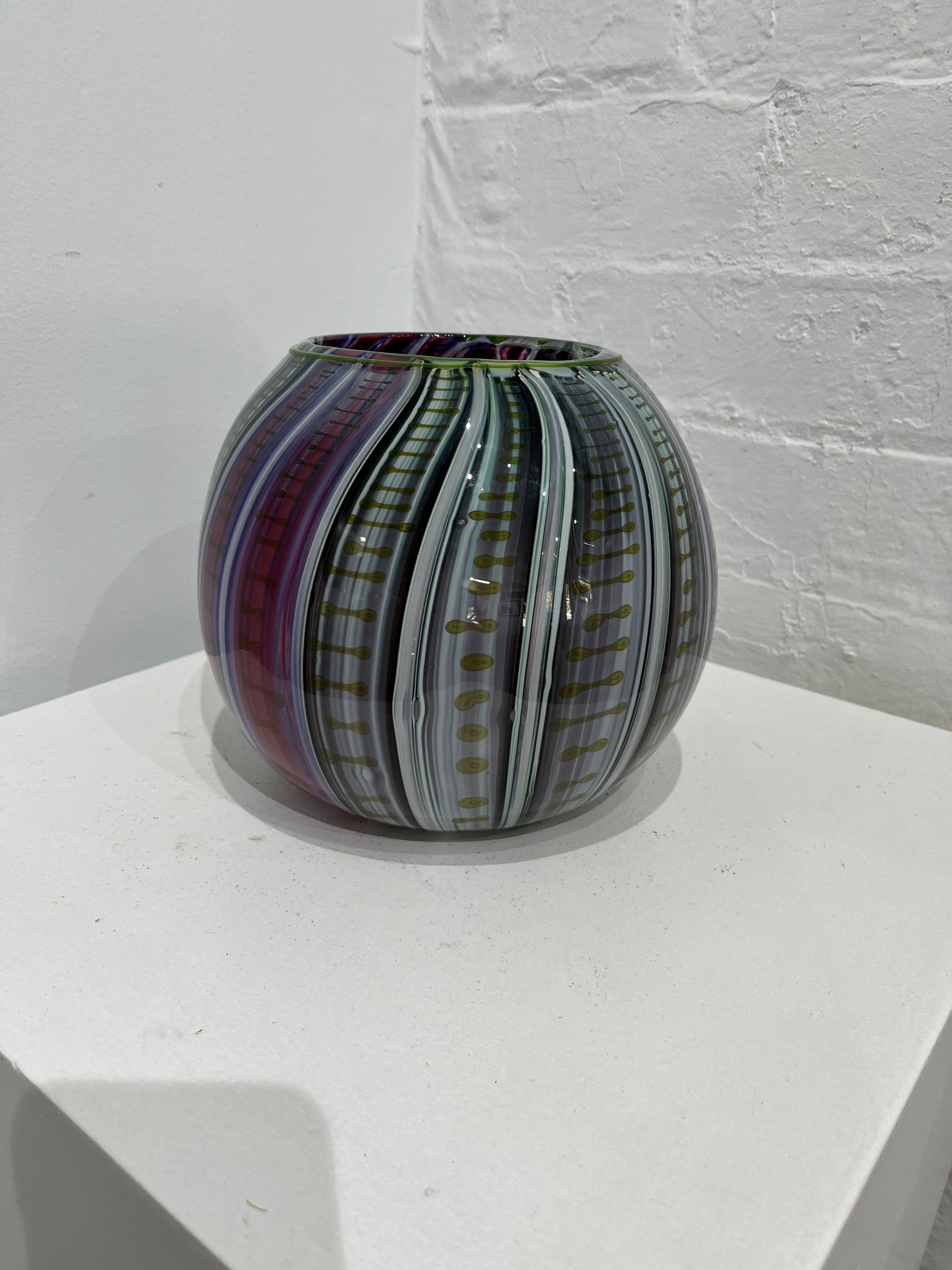 A hand-blown glass vase with a striped pattern of purple, green, gray, and gold, displayed on a white geometric platform against a white brick wall.