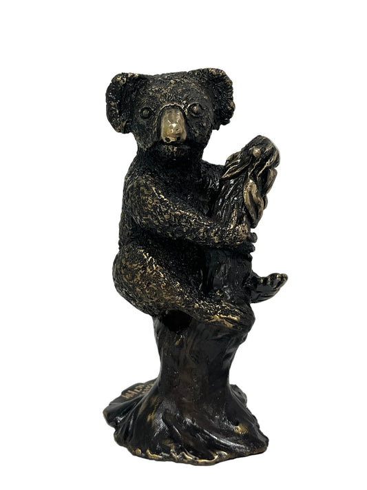 A dark bronze-finished sculpture of a koala clinging to a tree trunk, with metallic gold highlights on its face, paws, and trunk base, set against a plain white wall and surface.