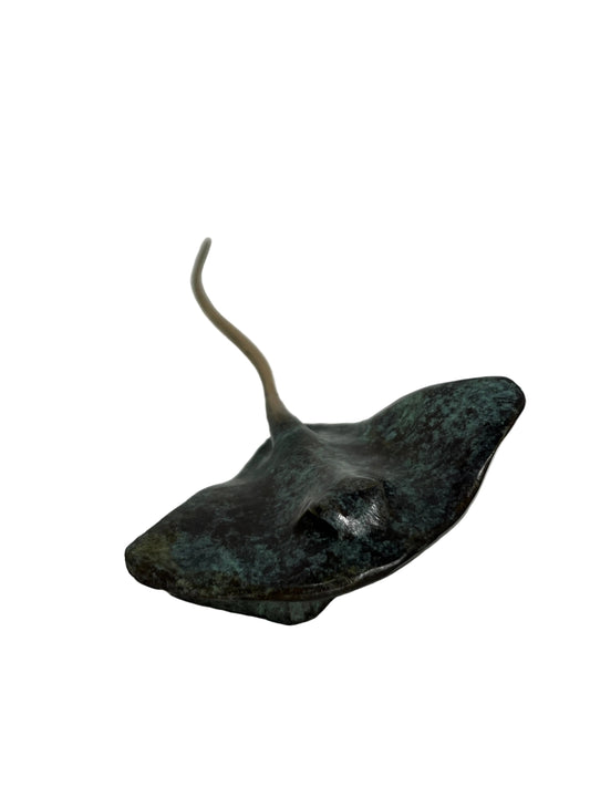 A small sculptural figurine of a stingray with a dark, textured surface showing greenish patina and a long, slender tail, photographed on a plain white surface against a white background.