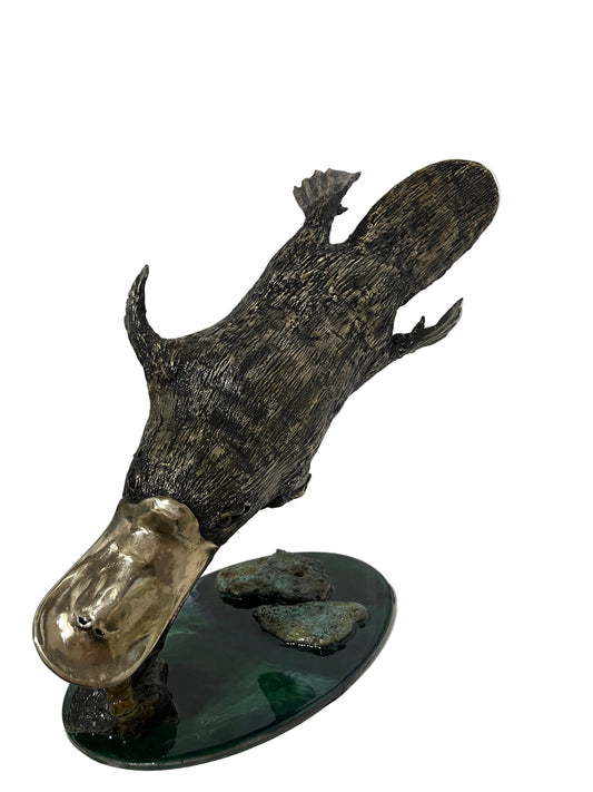 A platypus sculpture with a textured dark brown and black body, a metallic gold beak, and a dark green oval base with two faux rocks, displayed on a white shelf.