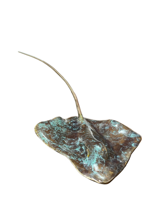 A leaf-shaped decorative sculpture with a weathered bronze and turquoise patina finish, placed on a wooden table in front of a framed abstract print.