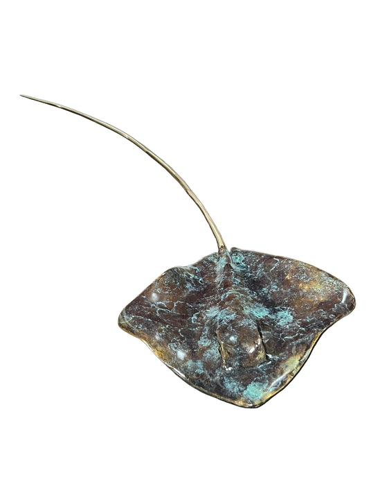A bronze sculpture of a stingray with a long, slender tail, displayed on a rustic wooden table.