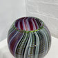 A spherical glass bowl with a marbled swirl pattern of purple, green, gray, and white, resting on a white surface against a white brick wall.