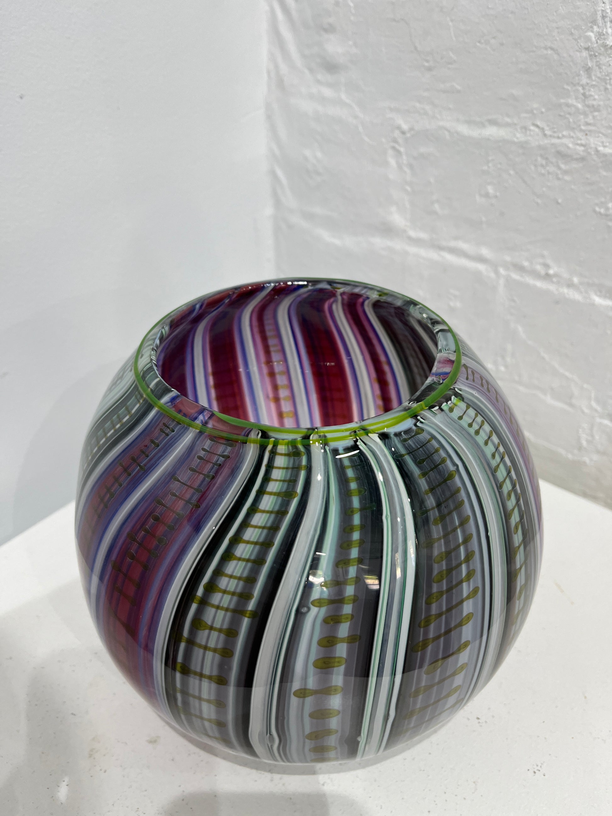 A spherical glass bowl with a marbled swirl pattern of purple, green, gray, and white, resting on a white surface against a white brick wall.