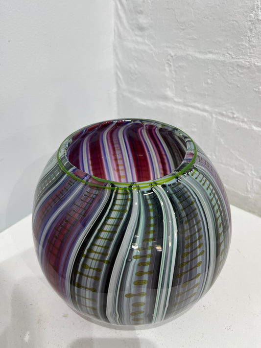 A spherical glass bowl with a marbled swirl pattern of purple, green, gray, and white, resting on a white surface against a white brick wall.
