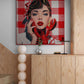 A large square canvas print featuring a stylized pop-art portrait of a woman with red lips and a red-and-white checkered background, displayed on a wall above a light wood cabinet in a modern interior setting.