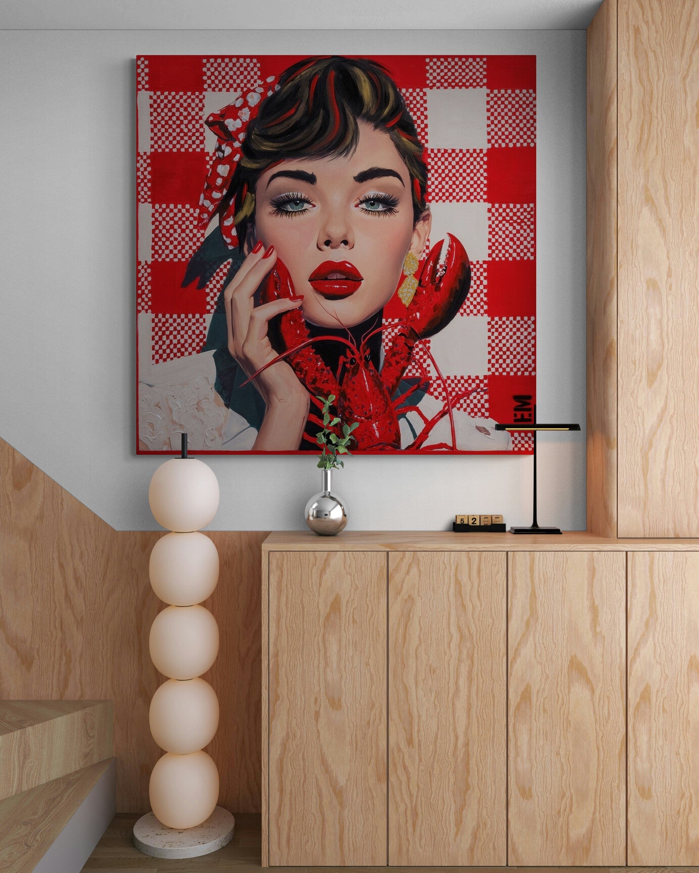 A large square canvas print featuring a stylized pop-art portrait of a woman with red lips and a red-and-white checkered background, displayed on a wall above a light wood cabinet in a modern interior setting.