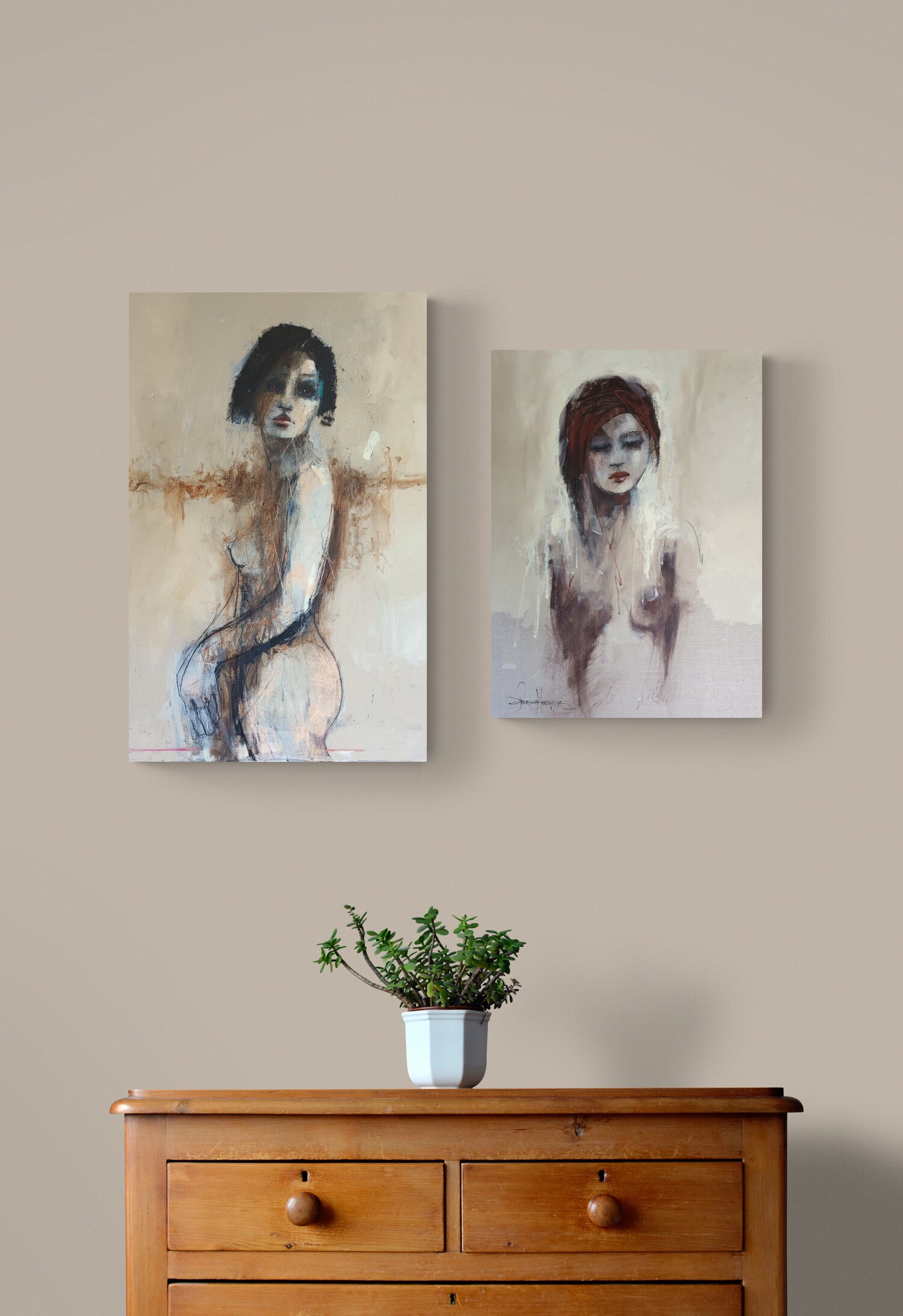 Two vertical canvas art prints depicting abstract female figures, displayed side-by-side on a beige wall above a wooden chest of drawers with a small potted plant.