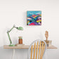 A colorful abstract landscape painting hanging on a white wall above a wooden desk with a green desk lamp, books, and a wooden chair.