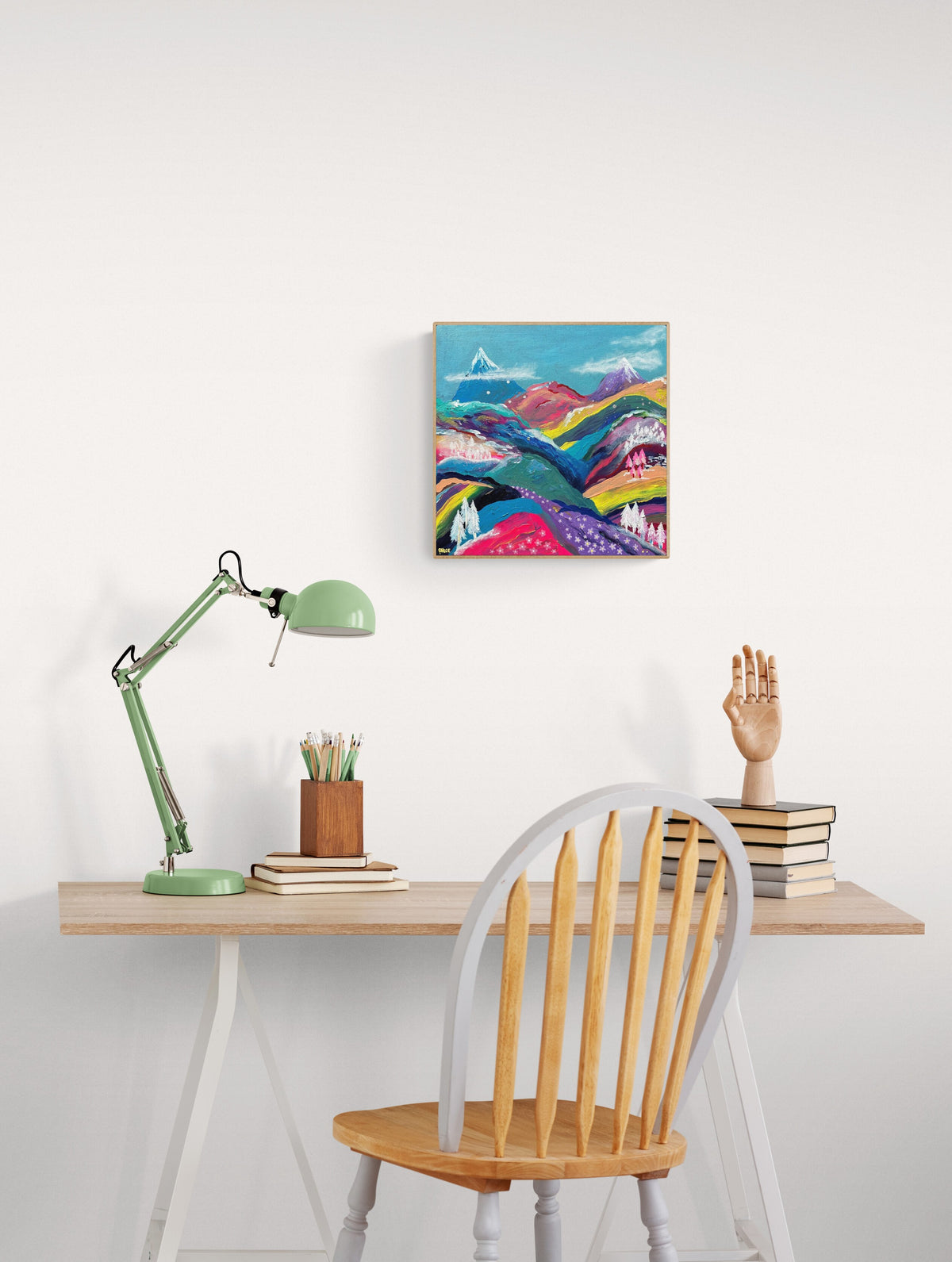 A colorful abstract landscape painting hanging on a white wall above a wooden desk with a green desk lamp, books, and a wooden chair.
